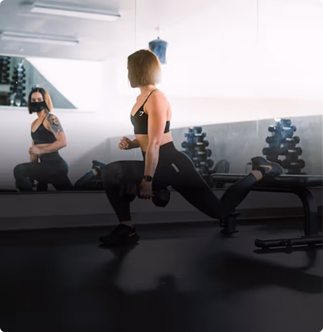 Woman in sportswear doing split squat with dumbbell in gym, her reflection visible in mirror.