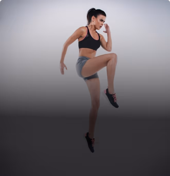Fit woman in black sports bra and gray shorts jumping with one knee raised during workout.