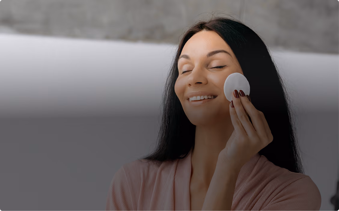 Smiling woman with long dark hair using a cotton pad on her cheek with eyes closed.