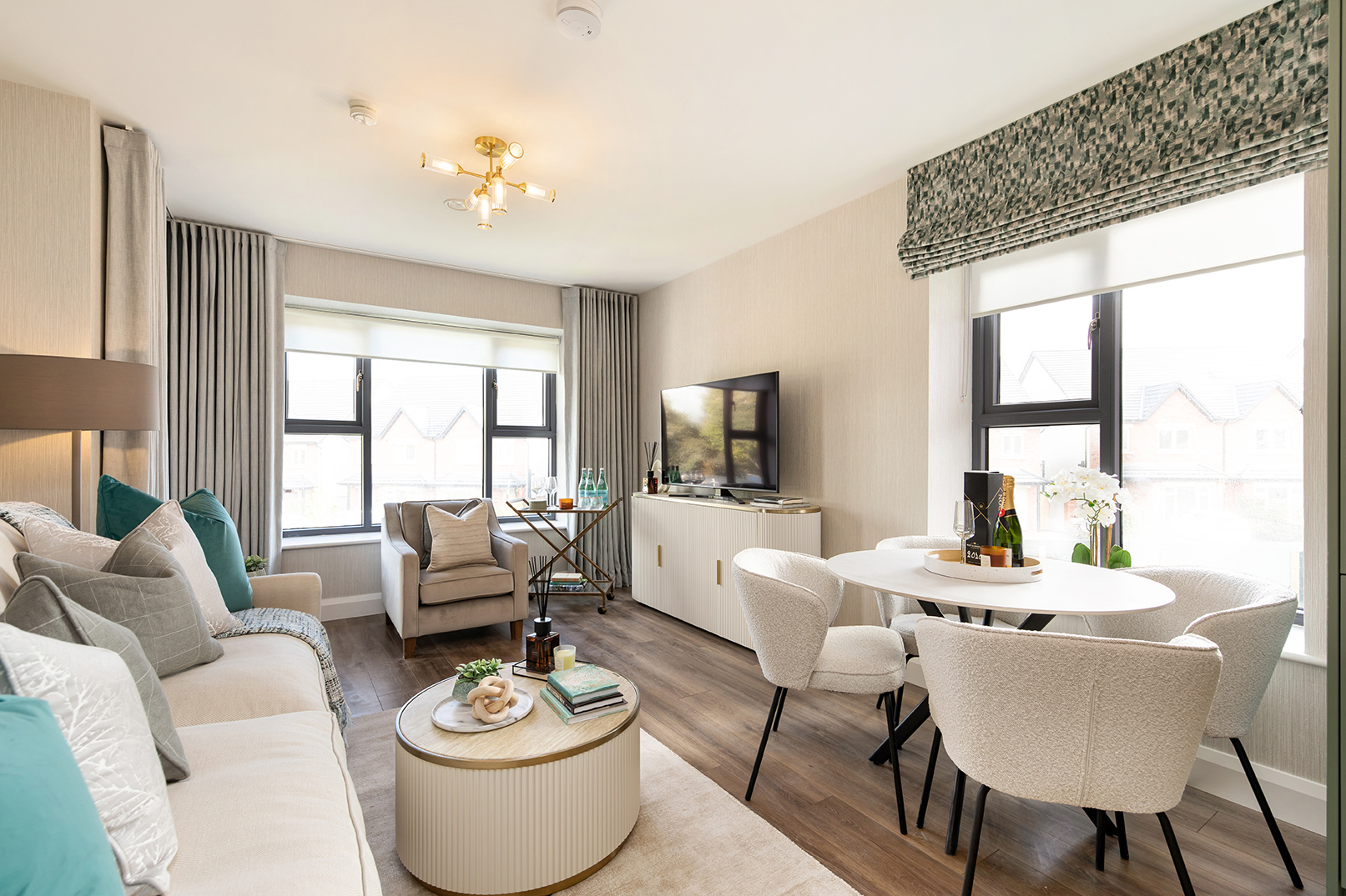 Bright modern living room with beige sofa, armchair, round coffee table, TV on white cabinet, and round dining table with four cream chairs near large windows.