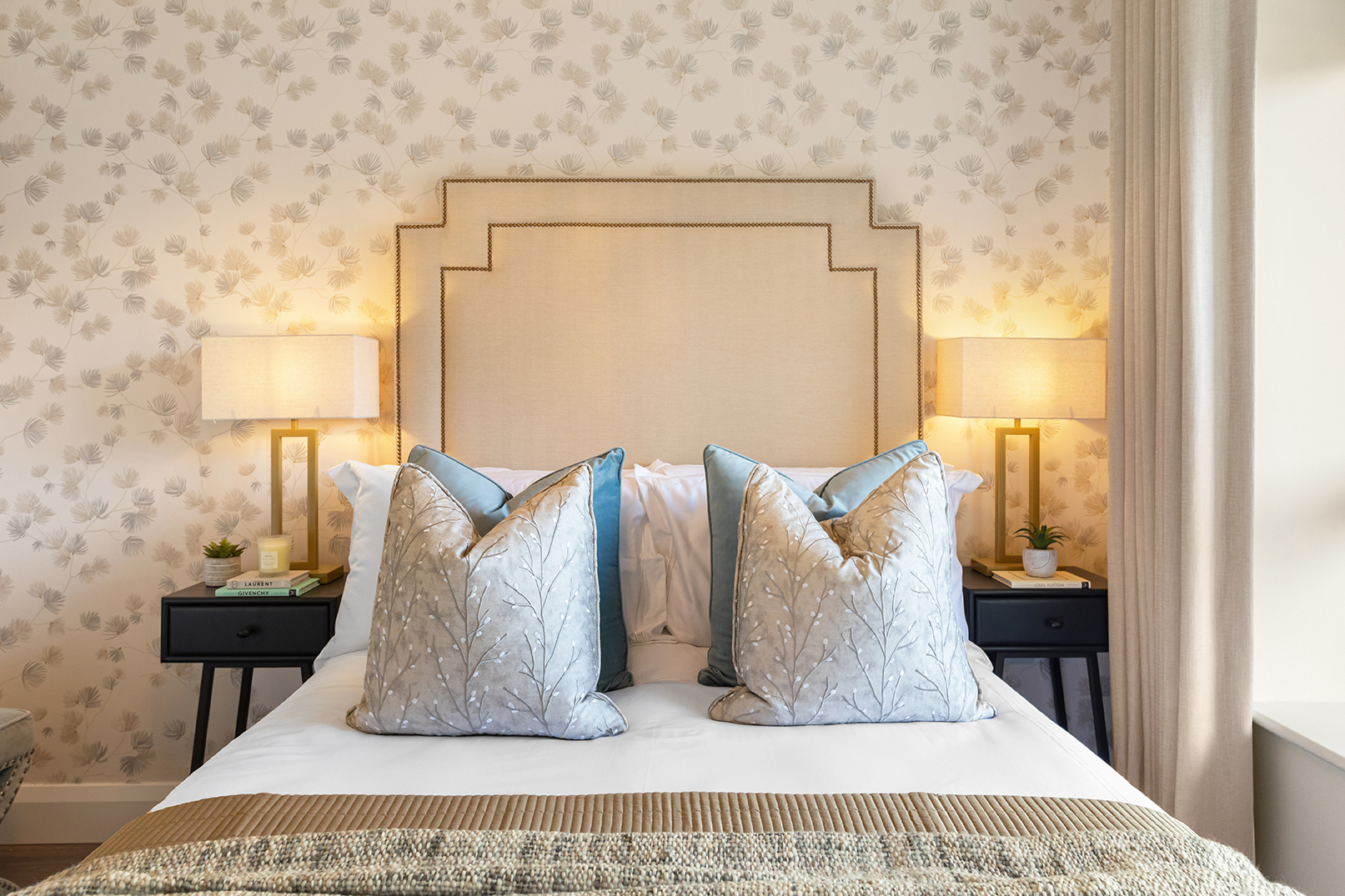 Neatly made bed with beige headboard, two decorative pillows with branch patterns, blue accent pillows, flanked by two nightstands each with a lit lamp and a small plant.