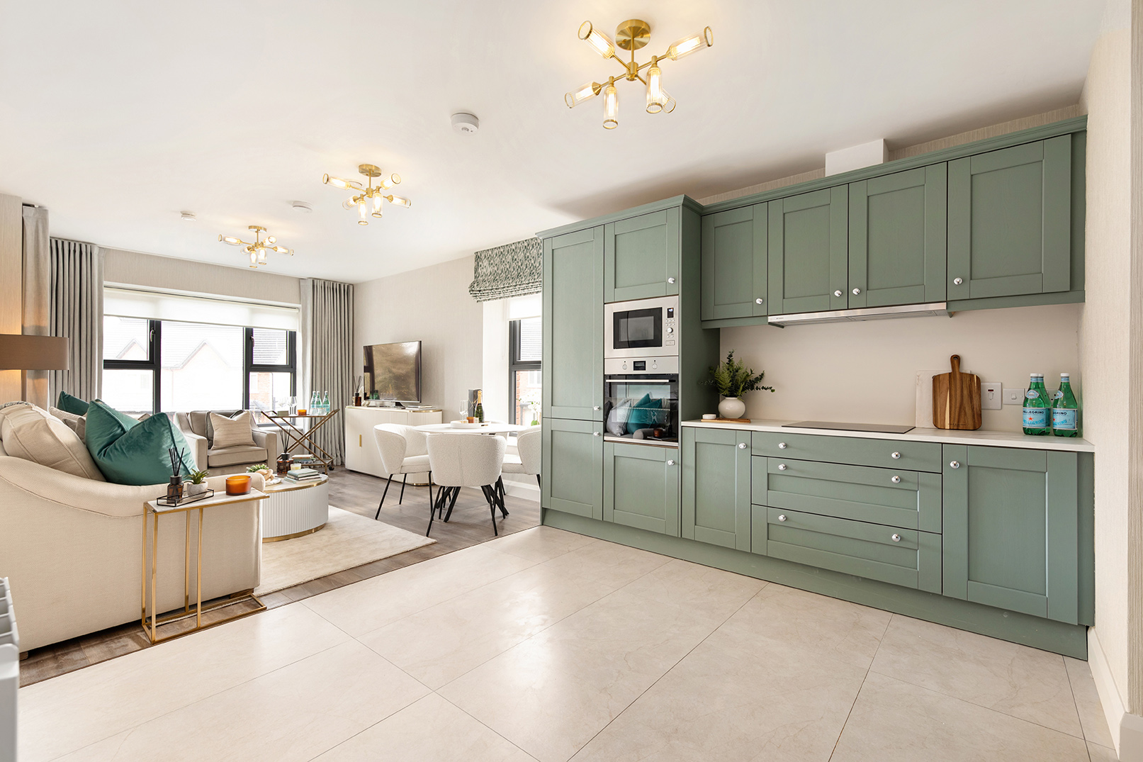 Open-plan modern kitchen and living room with muted green cabinets, beige tiled floor, cream sofa with green cushions, round coffee table, dining table with white chairs, and large windows with curtains.