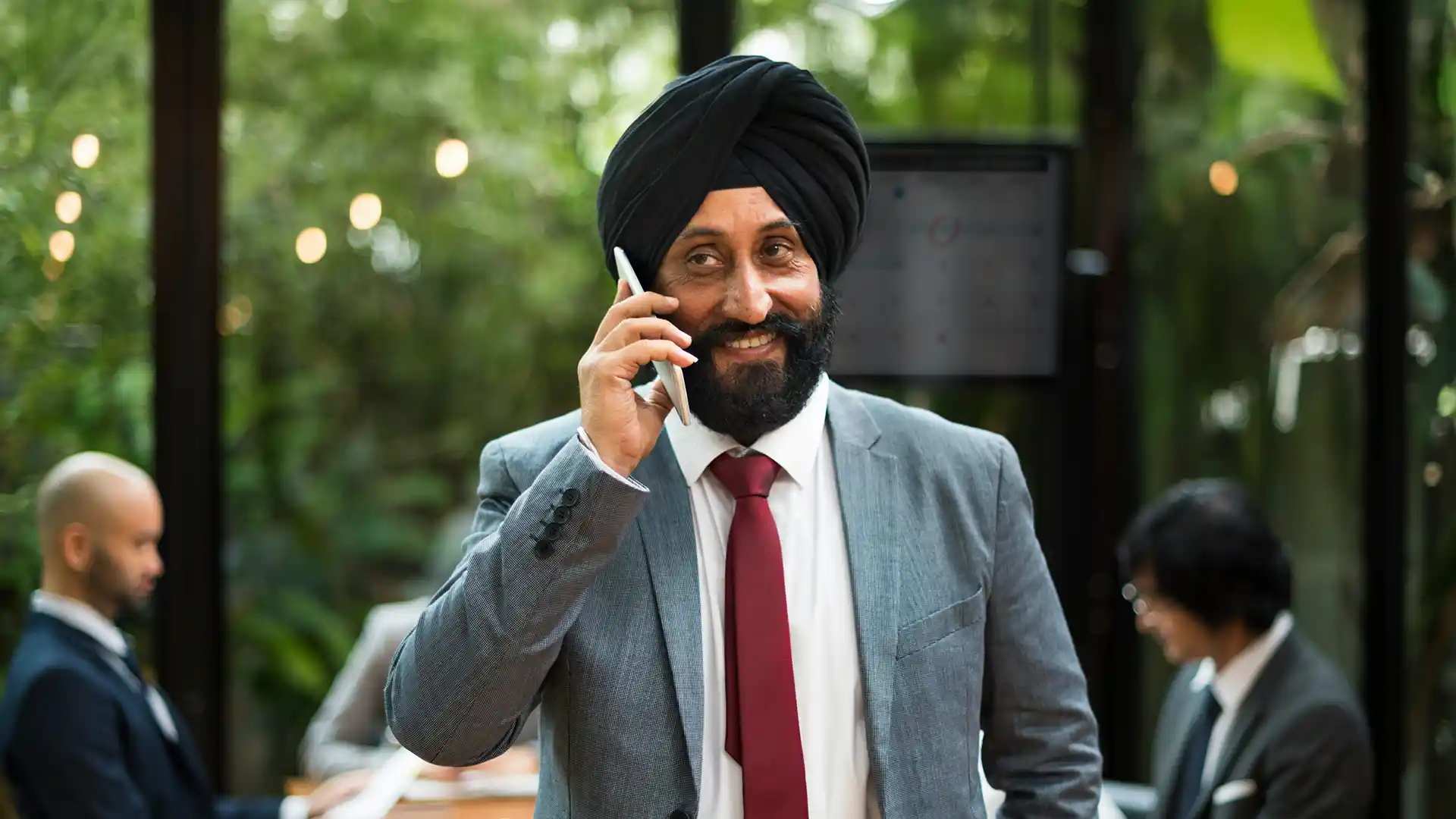 Person in a gray suit with a red tie and dark turban talking on a smartphone in an office setting with glass walls and greenery outside.