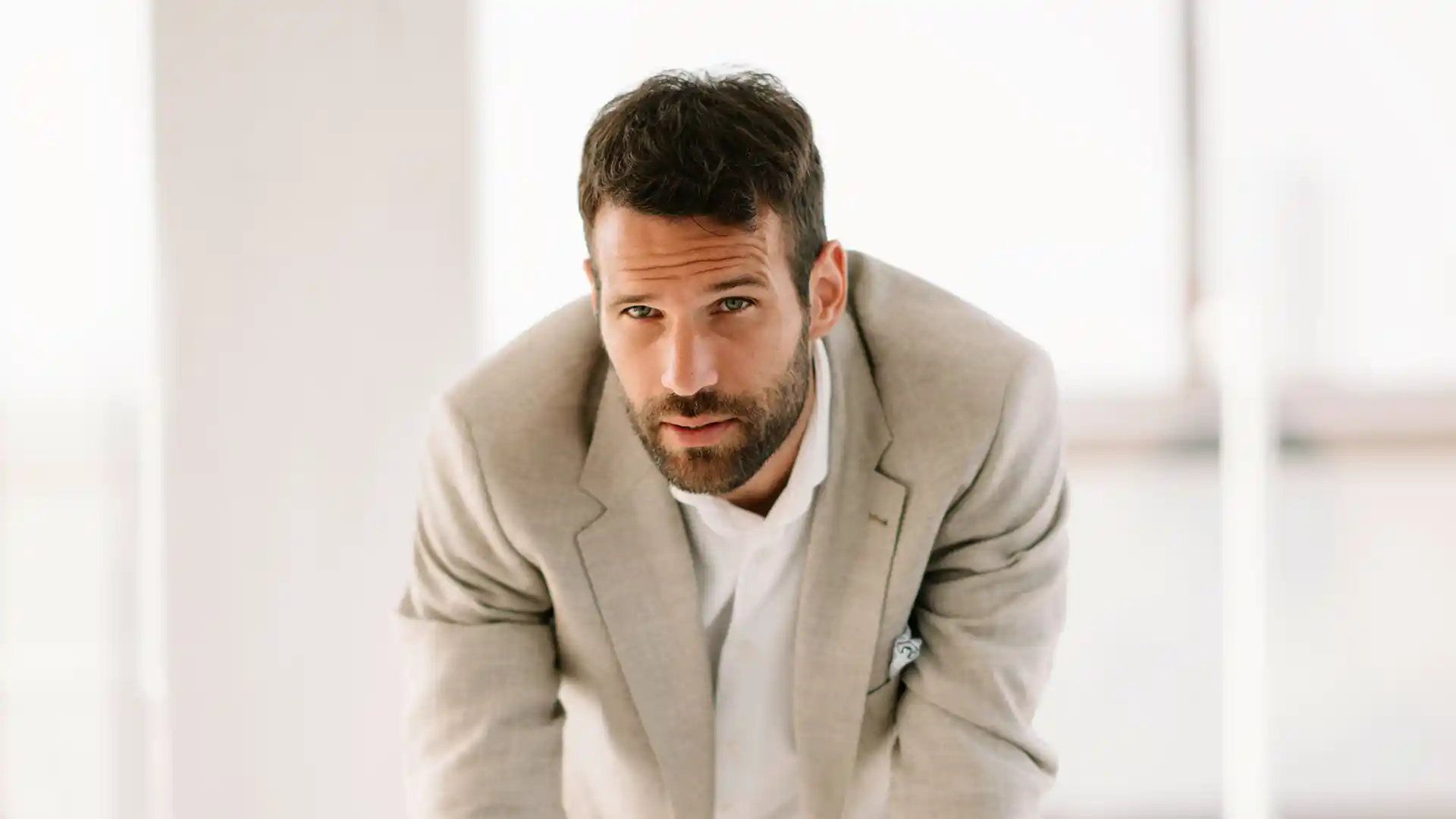  A close-up, centered portrait of a well-groomed man with a dark beard and short hair, wearing a light beige blazer and a white shirt.