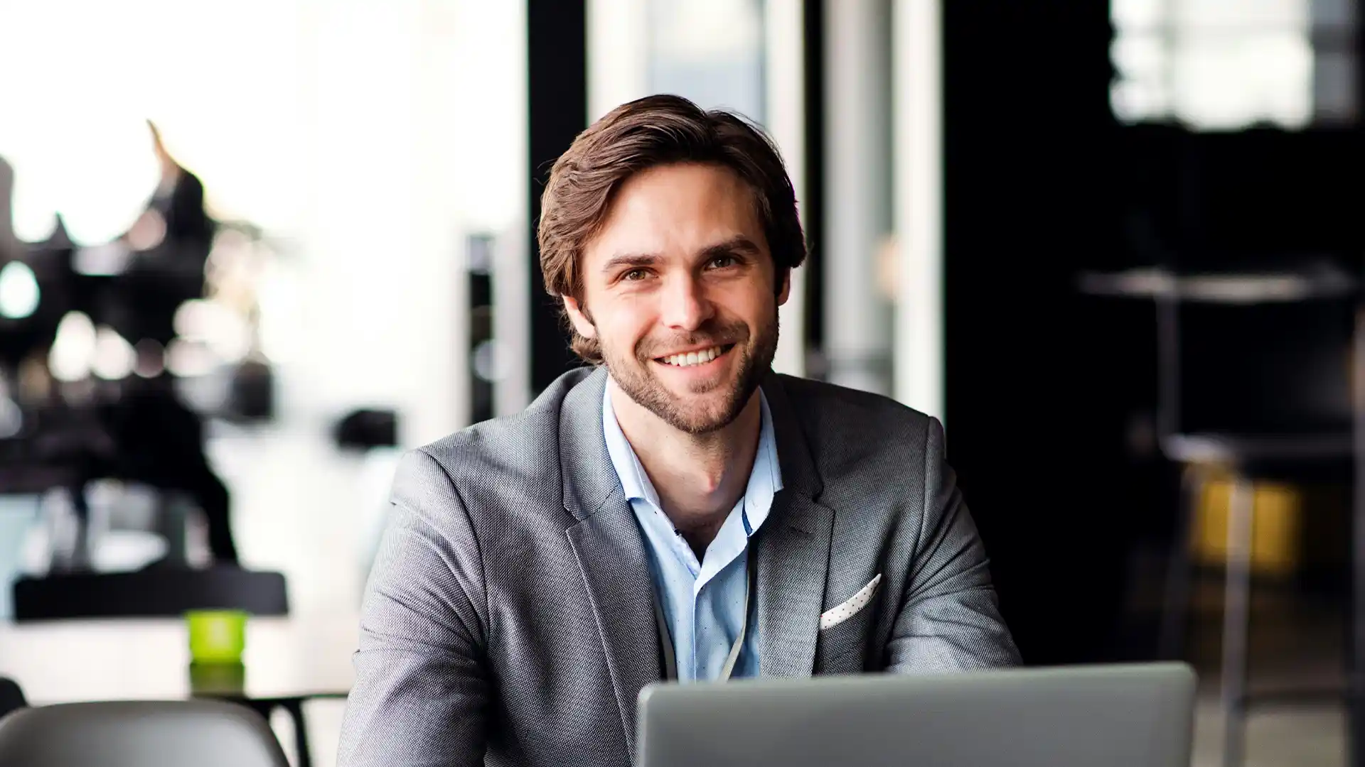 A happy, well-dressed man with brown hair and a beard smiles directly at the camera while sitting at a desk with a laptop in a modern office