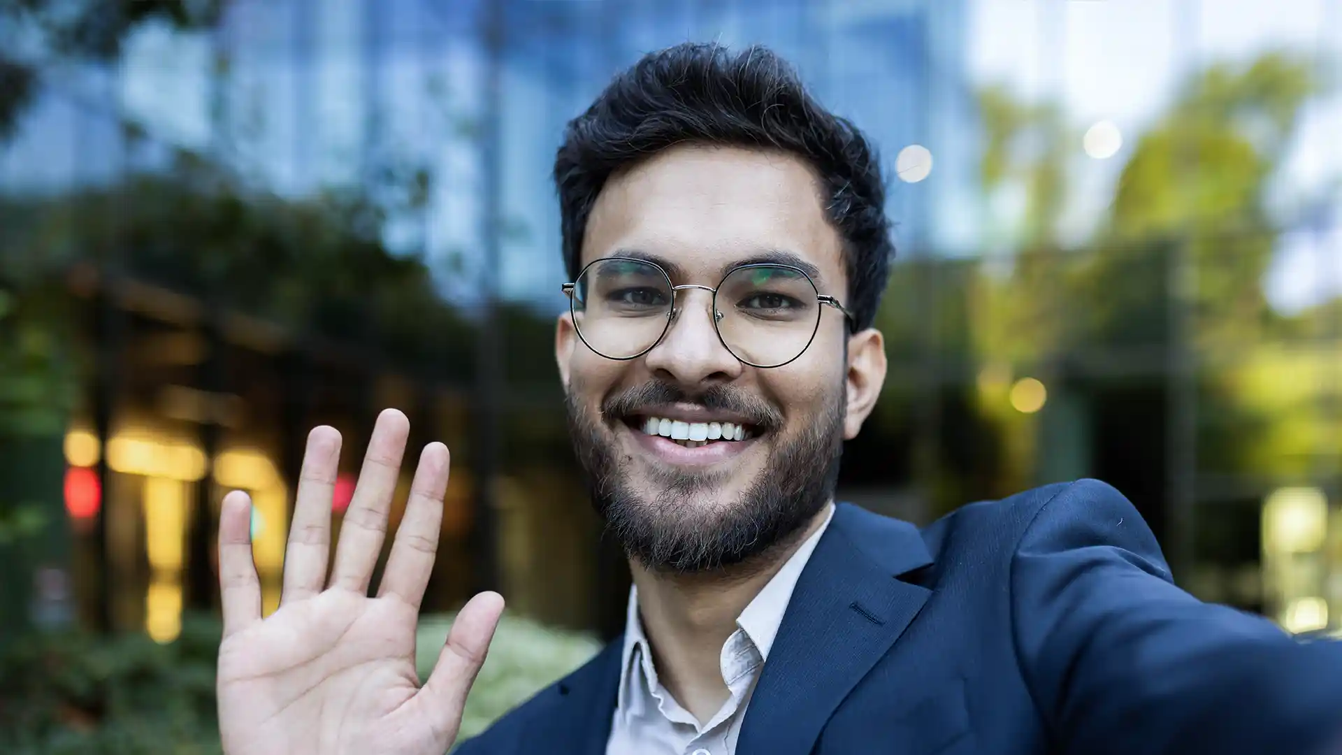 A young man with a beard and glasses smiles warmly while waving and taking a selfie outside a modern office building.