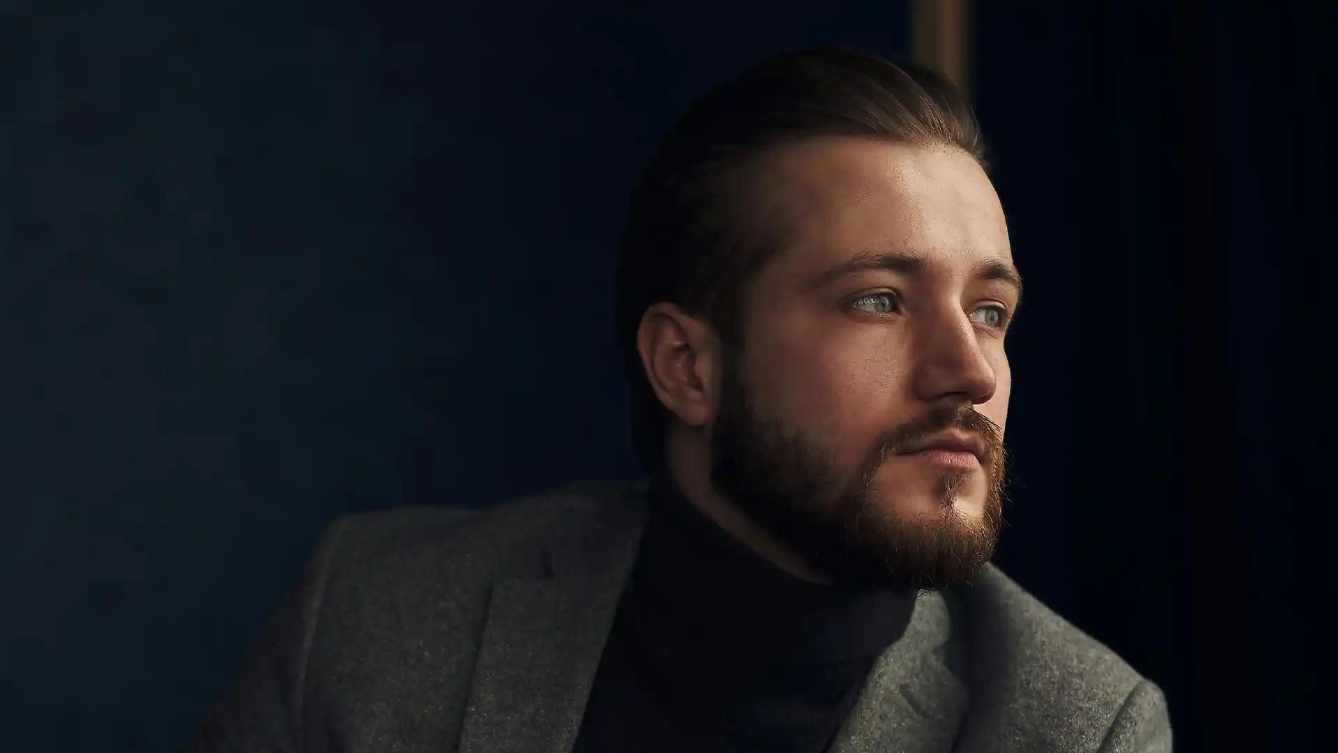 Person in a grey blazer and black turtleneck seated in a professional setting with dark background, representing business and financial