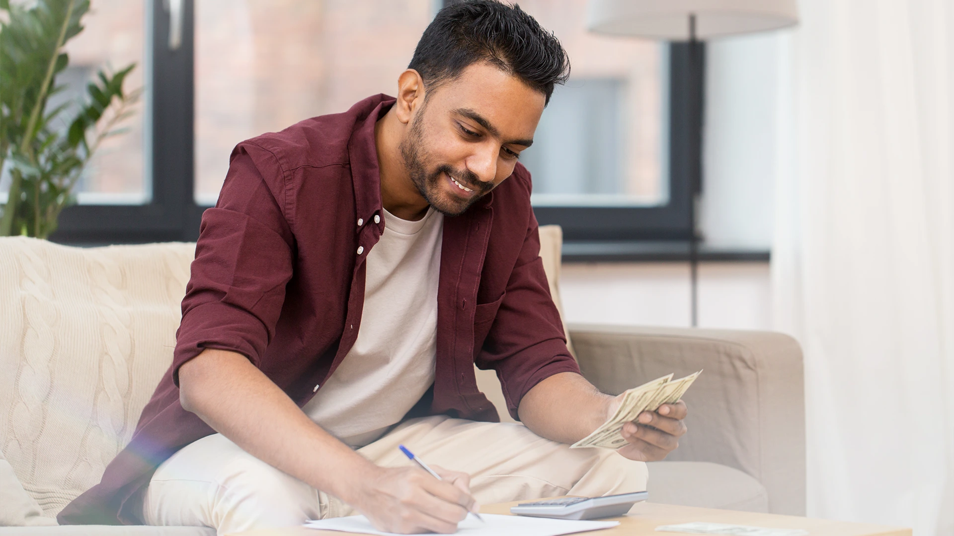 Person sitting on a couch counting cash and writing notes on a notepad, with documents and money spread on a table in a bright living room.
