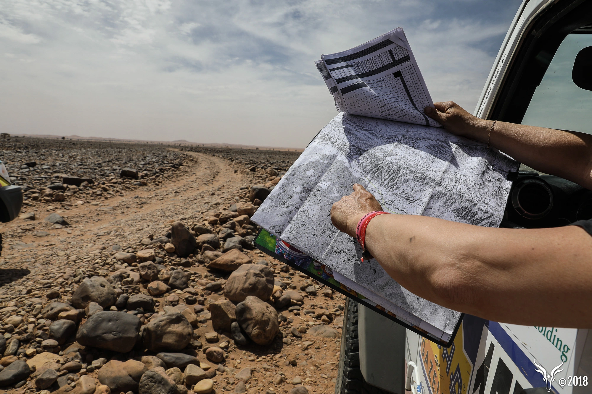 Un bras pointant du doigt un point sur une carte, par la fenêtre d'une voiture, dans le désert.