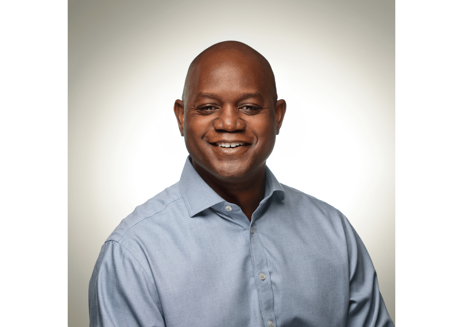 A man is looking straight ahead, smiling. He is wearing a light blue shirt and has a smart, business appearance. 