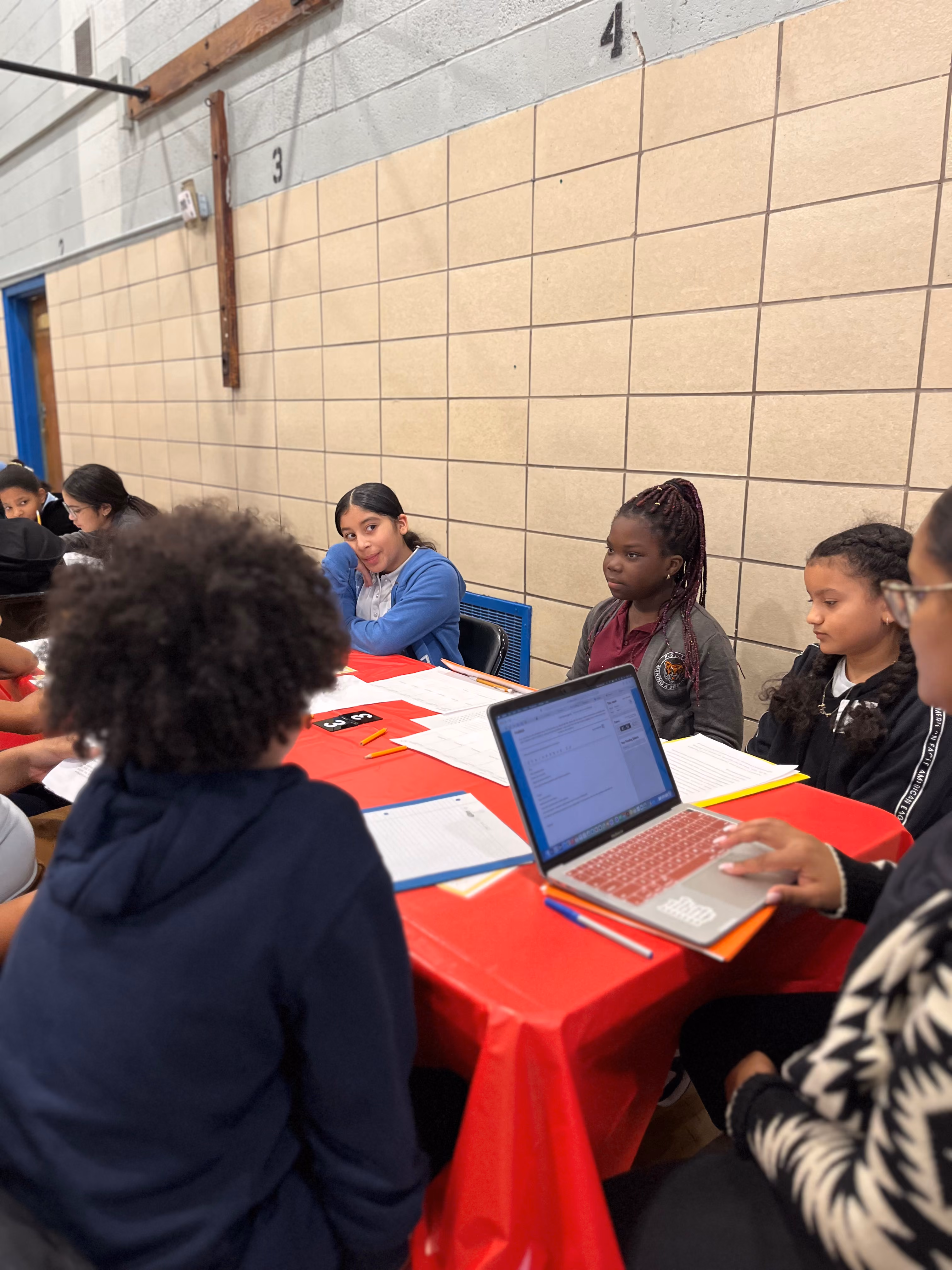 Students working at tables with laptops and papers in gymnasium