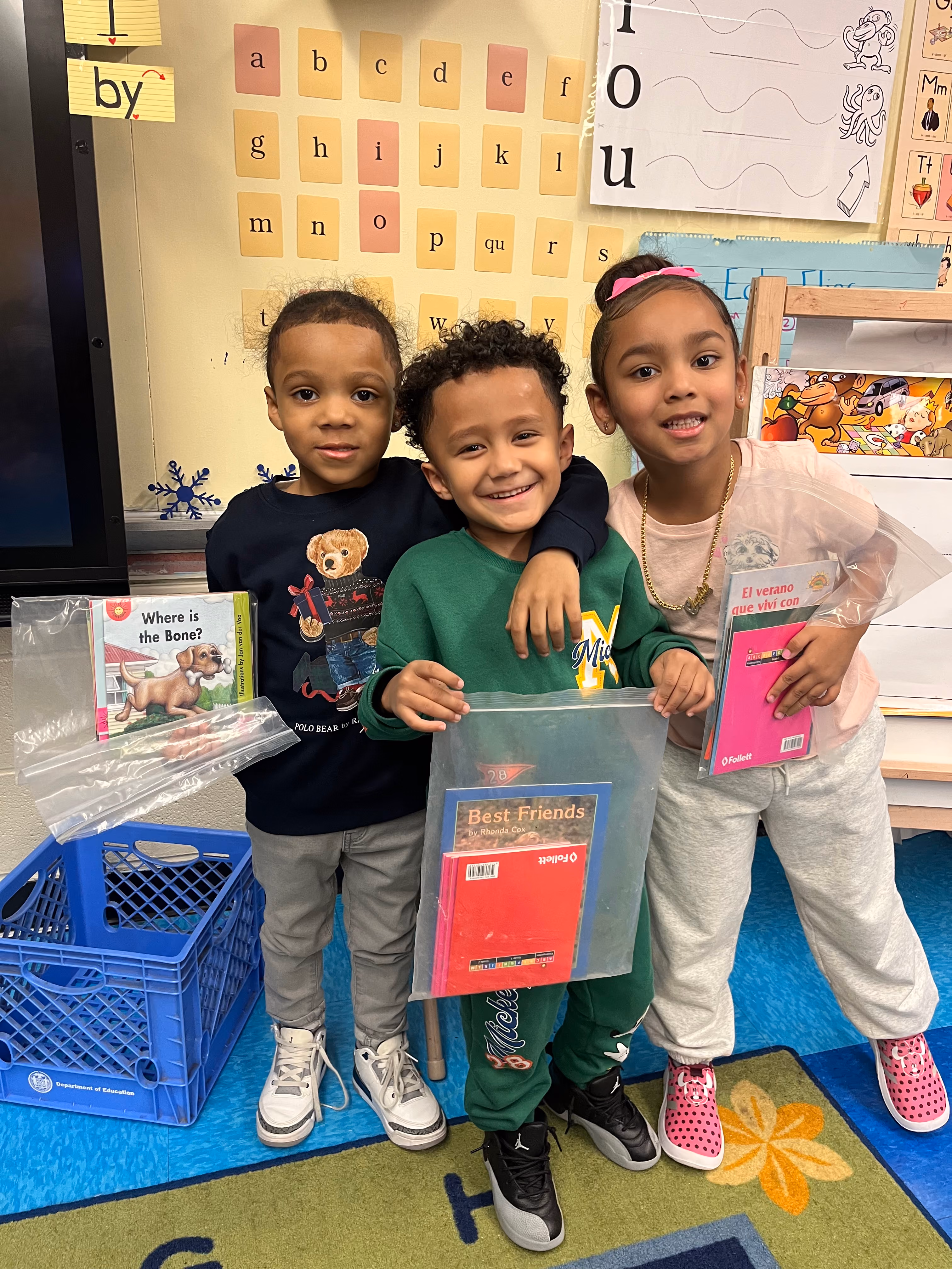 Three kindergarten students smiling and holding take-home book bags