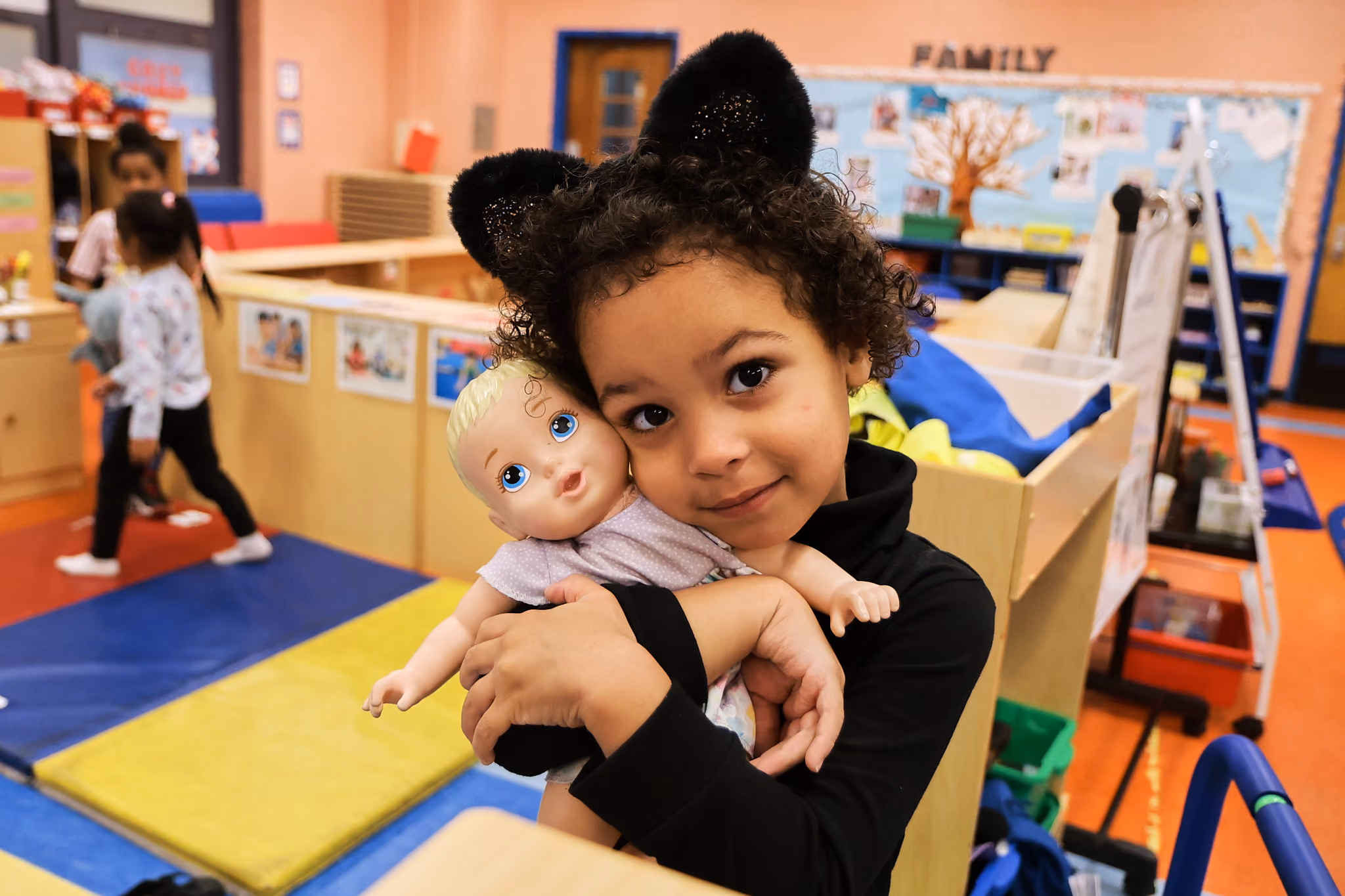 Young student in cat ears hugging a baby doll in classroom