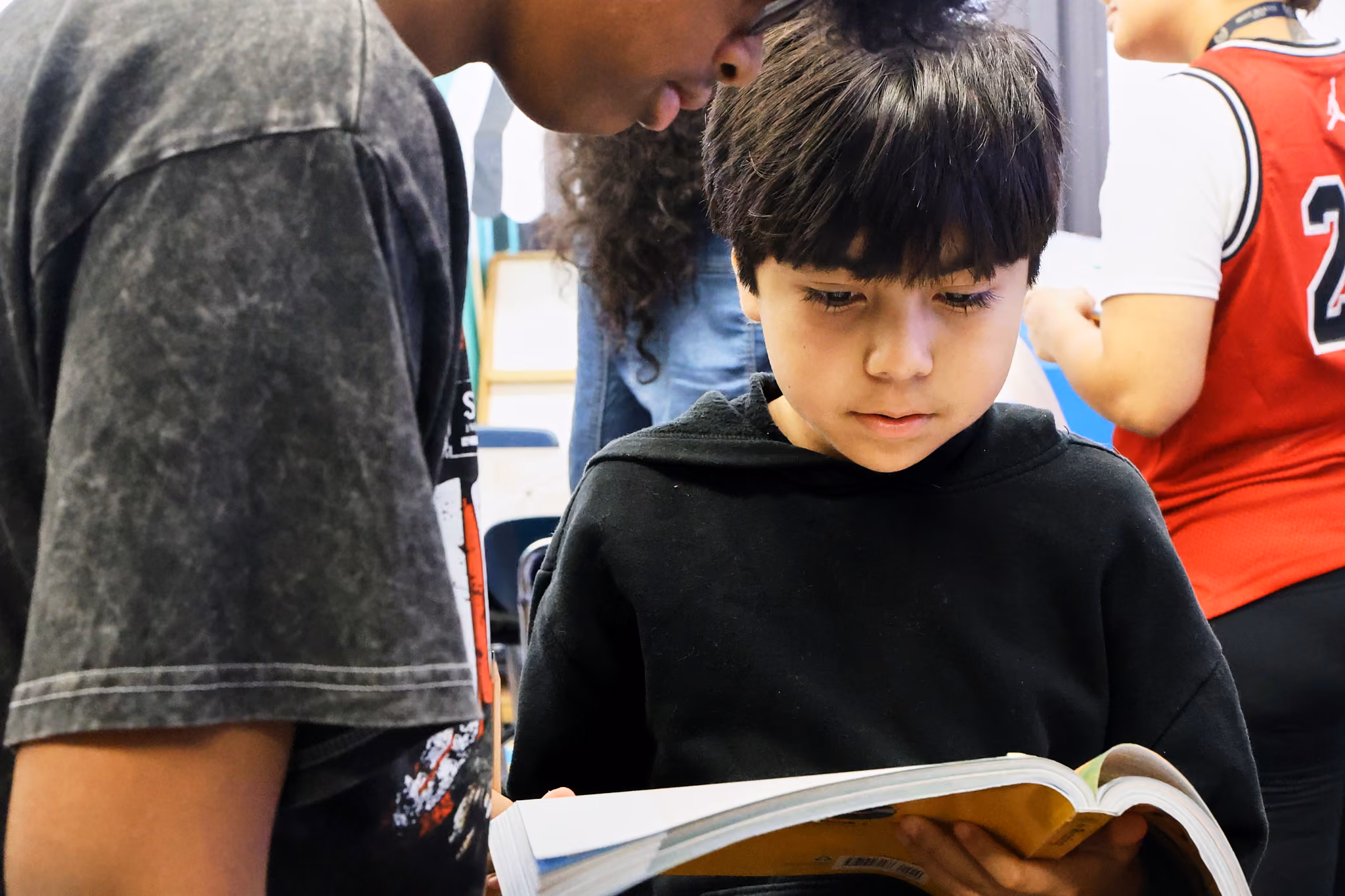 Student reading a book with classmates nearby in classroom
