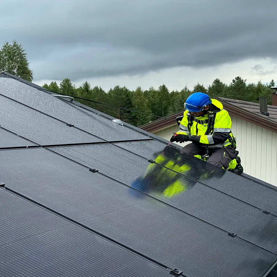 Työturvallisuusvarusteisiin pukeutunut työntekijä asentaa aurinkopaneeleja katon kaltevalla pinnalla pilvisenä päivänä.