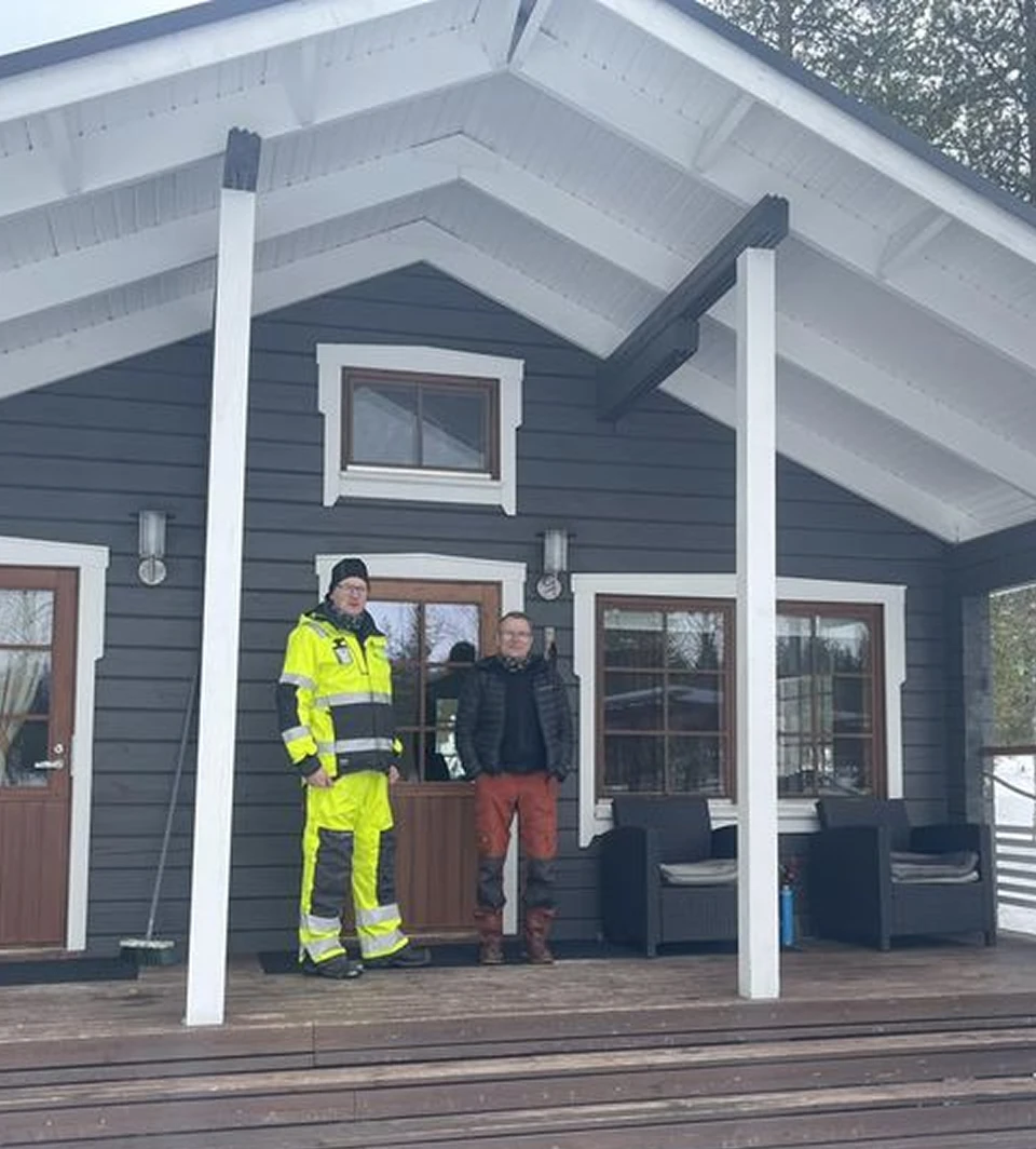 Asentaja ja toinen mies tumman puutalon kuistilla