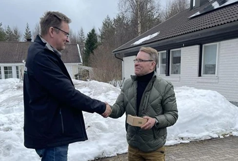 Kaksi miestä kättelee lumisella pihalla, toisella kädessään pieni laatikko