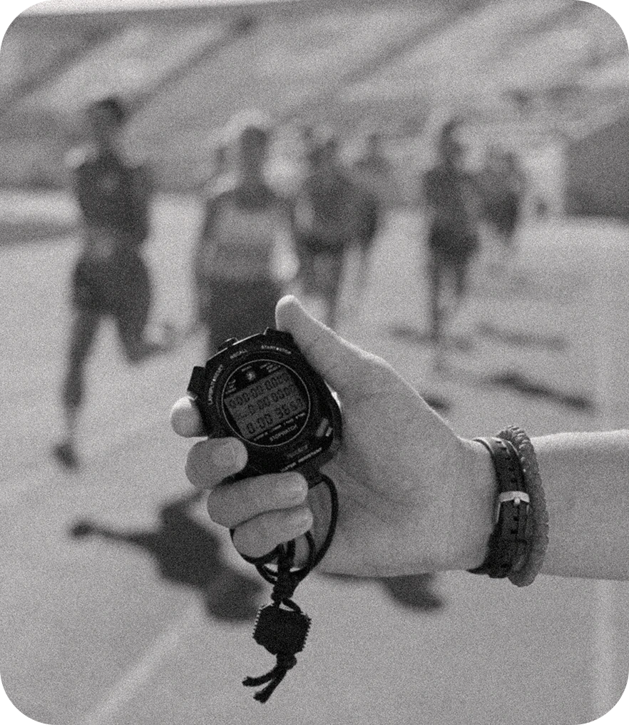 A coach holding a stopwatch trackside as runners approach on the track behind.