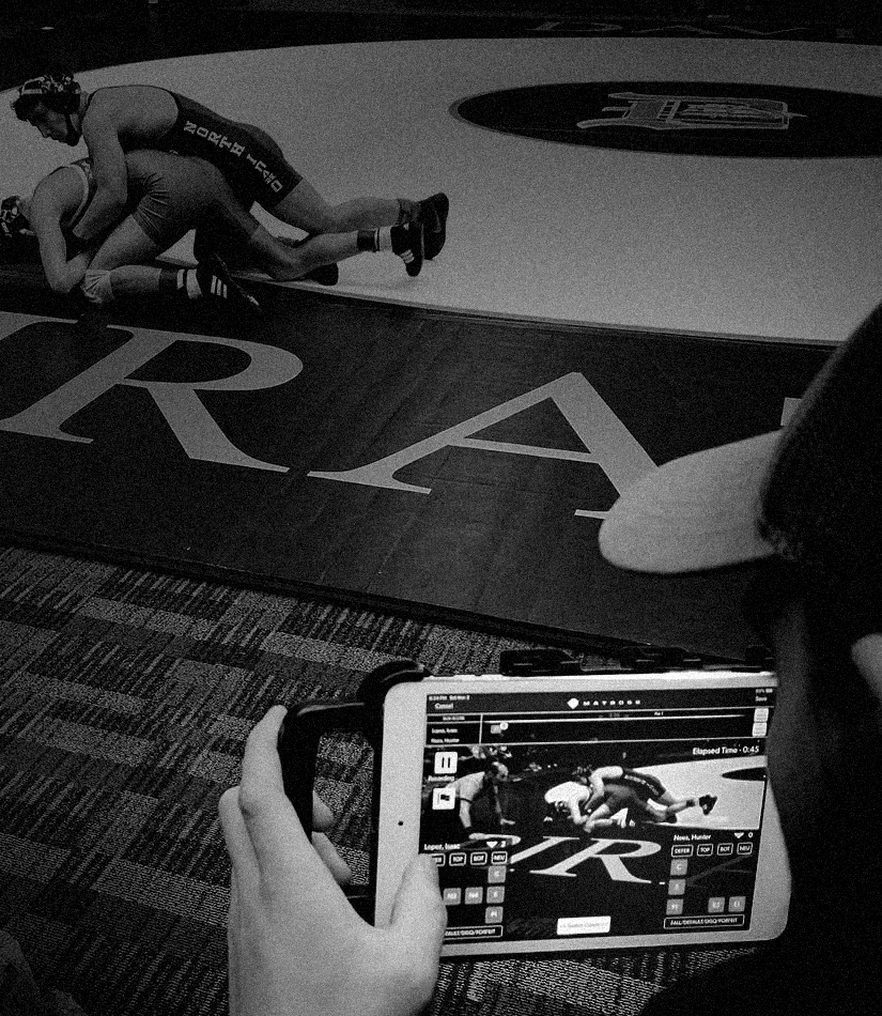A wrestling coach mat-side holding a tablet running MatBoss, recording and scoring the match taking place in front of him.