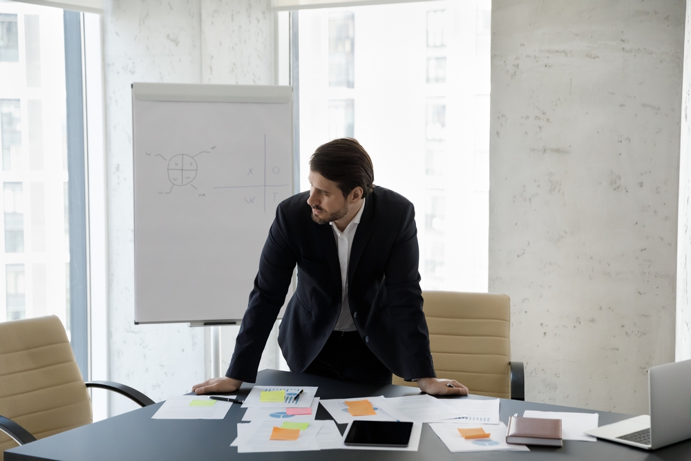 Frustrated Male-Presenting Executive Leaning over conference table looking to their right