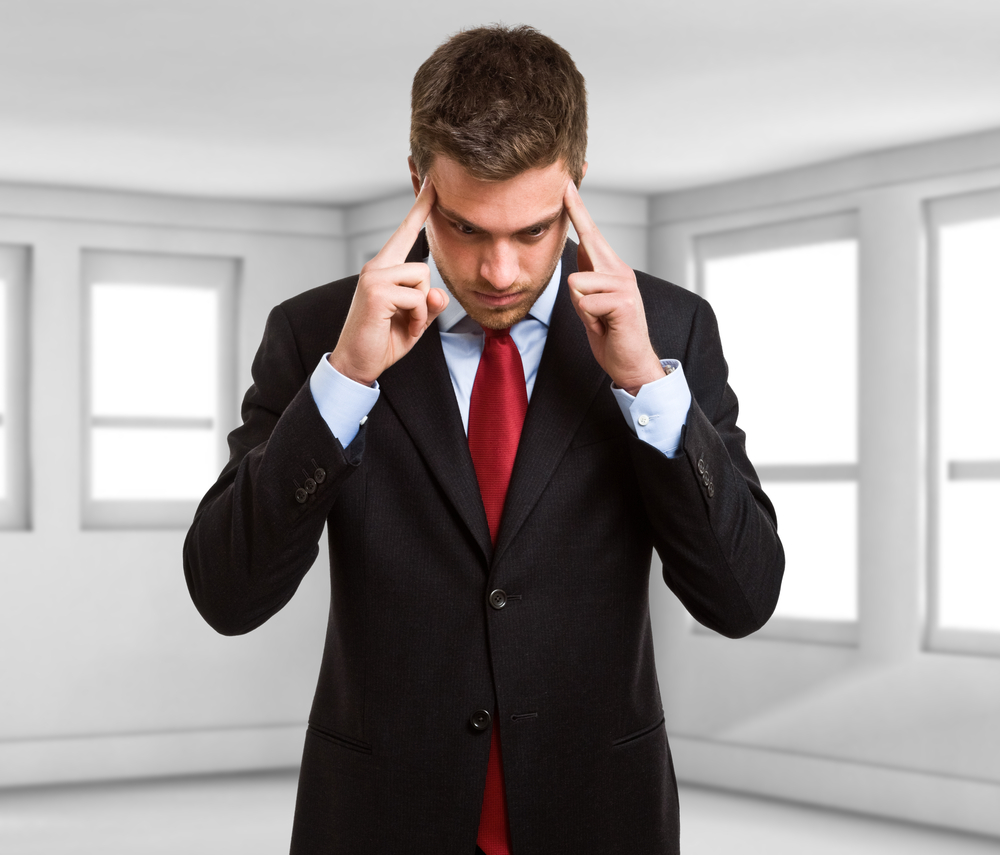 Young, male-presenting white person in a dark suit, white shirt and red tie, fingers to temple and concentrating