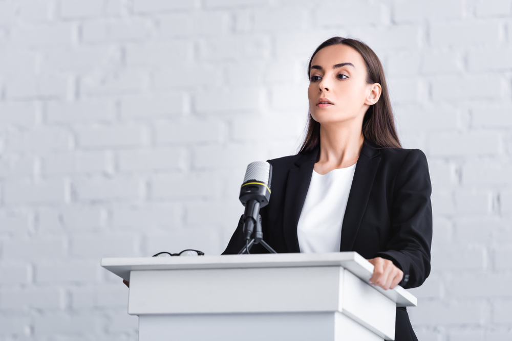 Young white women with straight brown hair gripping a podium hard with a concerned expression