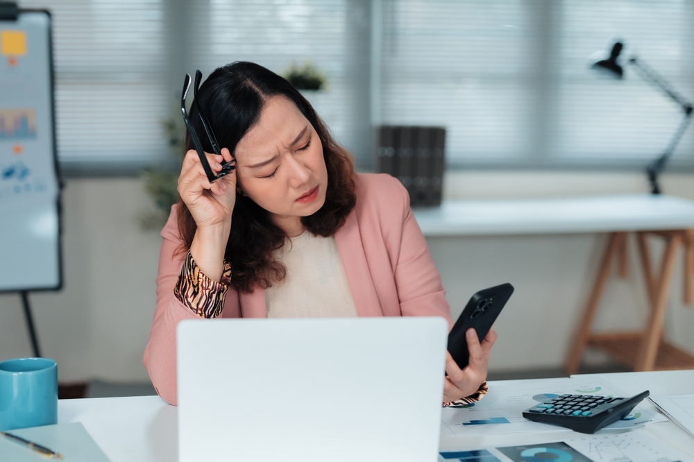 Asian Woman at desk looking tired and holding a cell phone