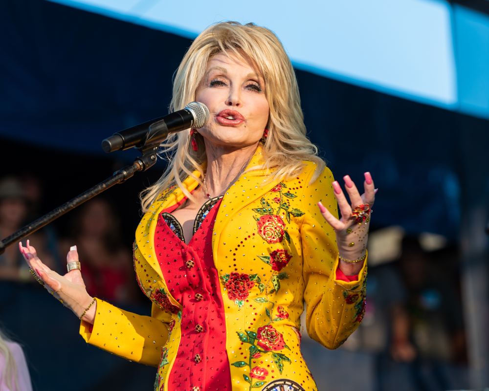 Dolly Parton at the mic wearing a yellow jacket over a red bustier