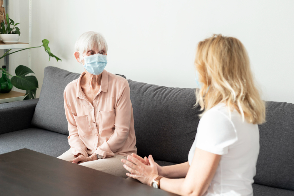 Elderly woman wearing mask speaks with younger woman on gray couch indoors