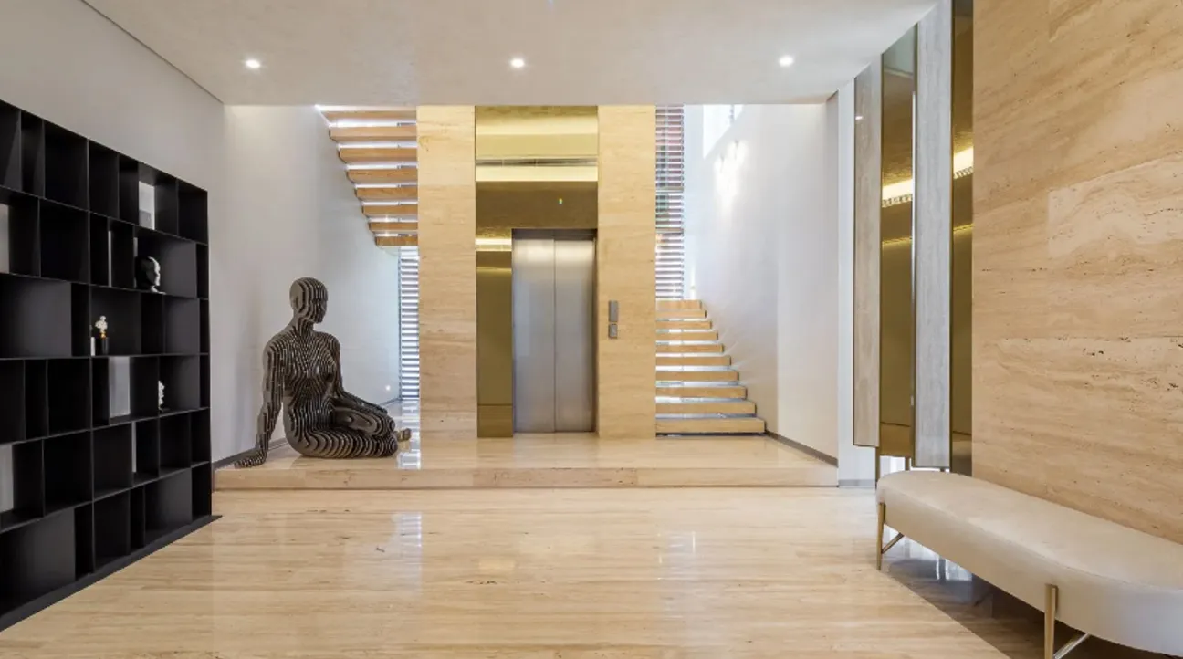 Contemporary hallway featuring a metal sculpture, elevator with gold details, and wooden stairs in a luxury villa.