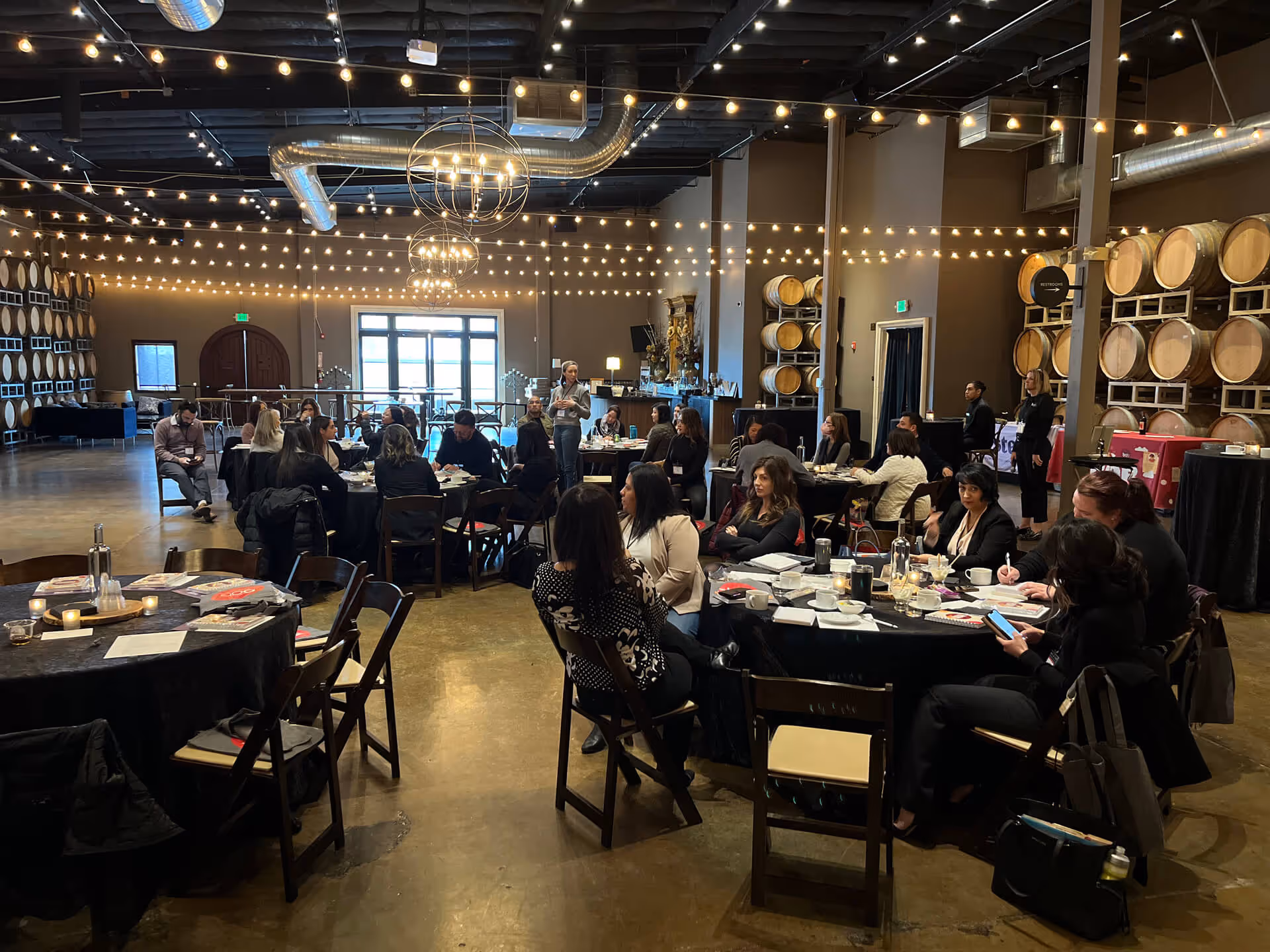 Attendees seated at tables in a winery event space with string lights