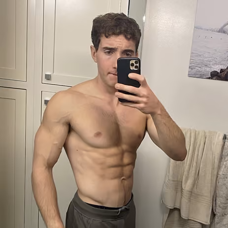 Shirtless man with muscular physique taking a mirror selfie with a smartphone in a bathroom.