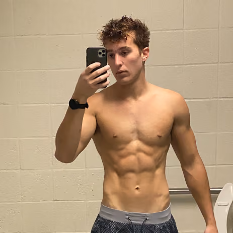 Young shirtless man with a fit muscular torso taking a selfie with a smartphone in a tiled bathroom.