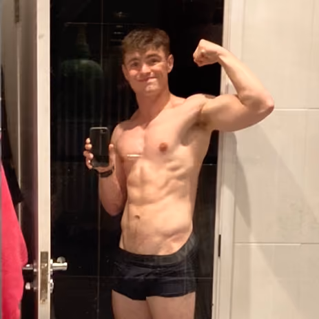 Young man wearing black shorts flexes his arm and takes a mirror selfie in a bathroom.