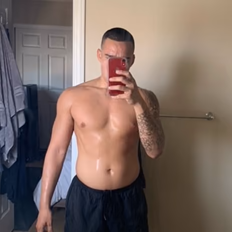 Shirtless man with a tattoo on his left arm taking a mirror selfie with a red phone in a bathroom.