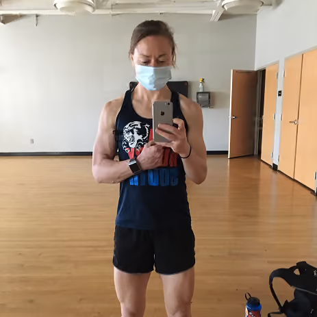 Person wearing a mask and workout clothes taking a mirror selfie in an empty gym room.