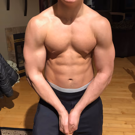 Shirtless muscular person flexing arms and showing toned chest and abs indoors on wooden floor.