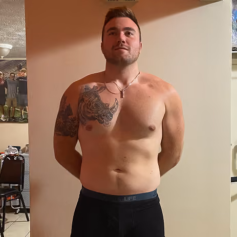 Shirtless man with a rhinoceros tattoo on right shoulder and arm stands against a beige wall.
