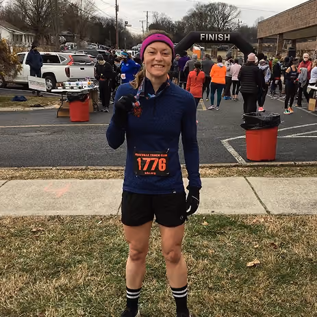 Runner holding medal and smiling near finish line at outdoor race event on a cloudy day.