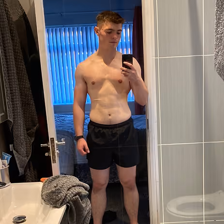 A shirtless young man wearing black shorts takes a mirror selfie in a bathroom.