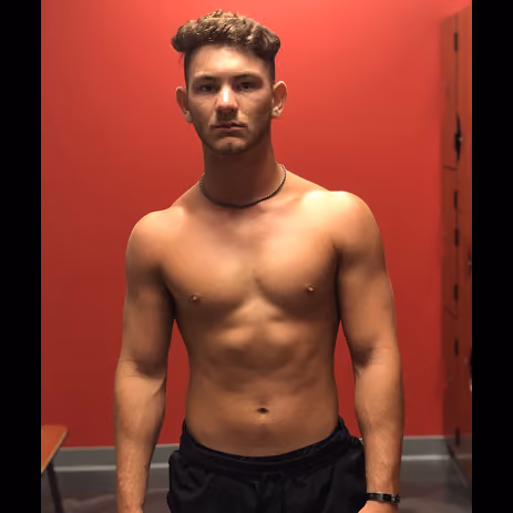 Shirtless man with muscular build standing against a red wall in a room.