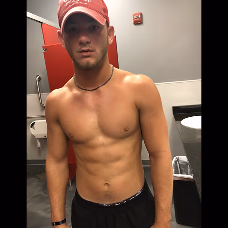 Shirtless young man wearing a red cap and black shorts standing in a bathroom with a red stall door.