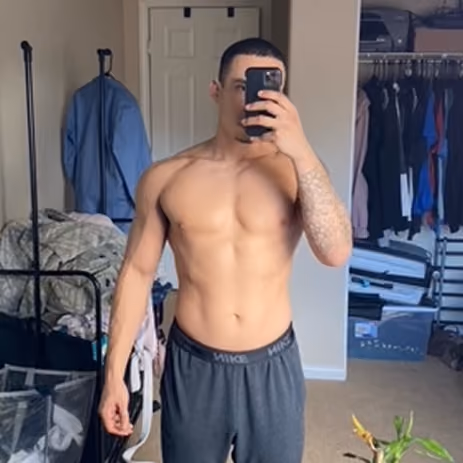 Shirtless man taking a mirror selfie in a bedroom with clothes and a closet in the background.