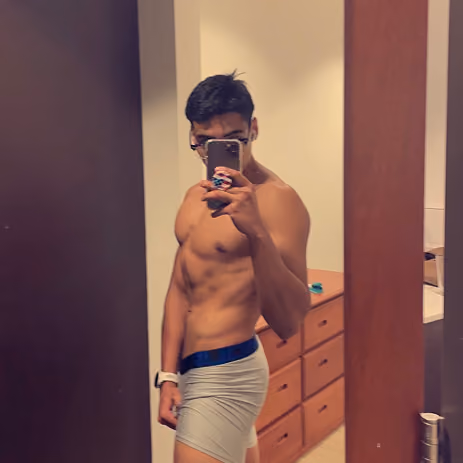 Muscular man taking a shirtless mirror selfie wearing light gray shorts in a bedroom.