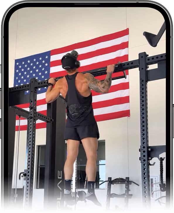 Muscular man doing pull-ups on a bar in a gym with an American flag hanging on the wall.