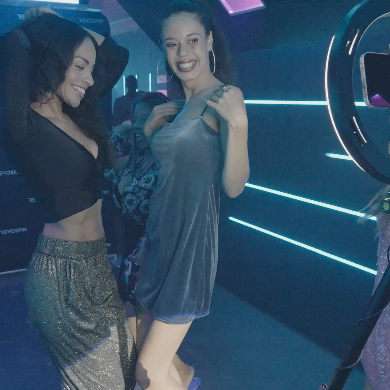 Two women smiling and posing next to a ring light in a neon-lit studio during a 360 booth moment.