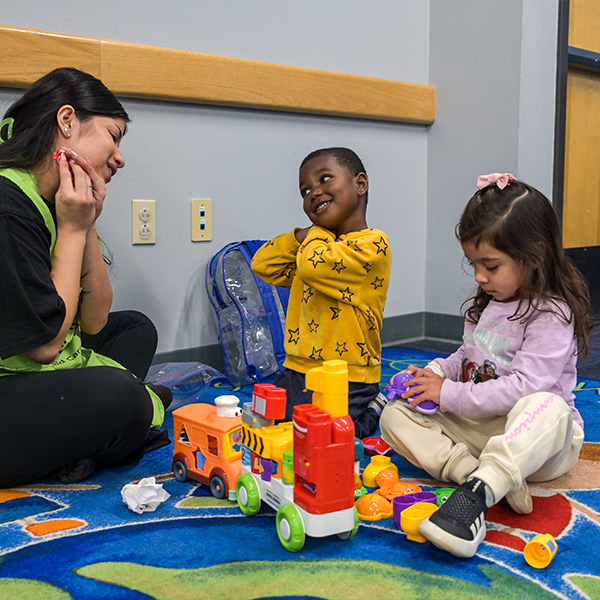 Young woman sitting on the floor with two children playing with colorful toys on a carpet in a classroom.