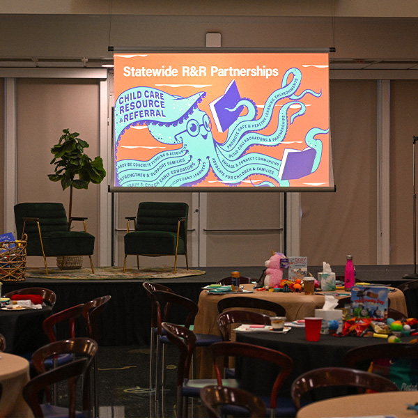 Conference room with round tables, chairs, and a stage featuring a large screen displaying an illustrated octopus promoting statewide child care resource and referral partnerships.
