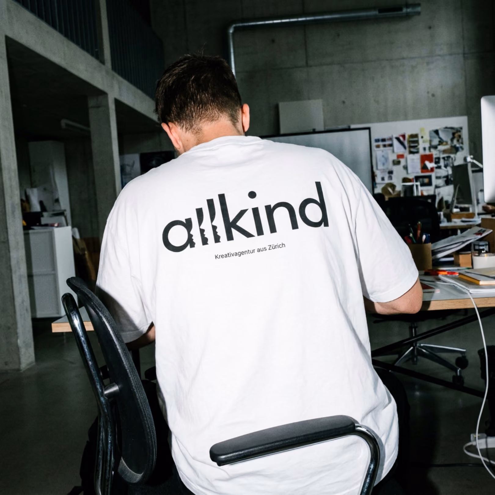 Person in white shirt with allkind logo and Schreiberei 'Kreativagentur aus Zürich' on back, sitting at desk.