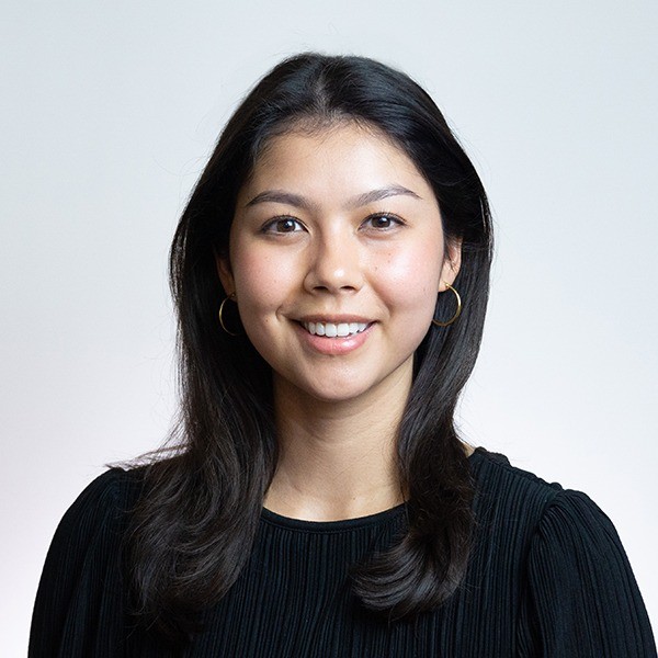 Portrait of a smiling young woman with long dark hair wearing hoop earrings and a black top against a light background.